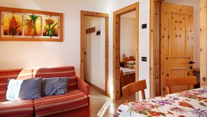 A room with a sofa, a dining table with a floral tablecloth, wooden doors, and a wall-mounted painting. - Pet-friendly Holiday Home Baita sulle Alpi with Mountain View, Garden, Terrace, BBQ & Wi-Fi (livigno)