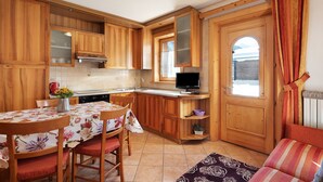 A compact kitchen with wooden cabinets, a dining table with chairs, a TV, and a view through a door. - Pet-friendly Holiday Home Baita sulle Alpi with Mountain View, Garden, Terrace, BBQ & Wi-Fi (livigno)