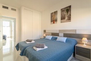 A hotel room with two beds, a wardrobe, a bedside table with a lamp, and artwork on the wall.