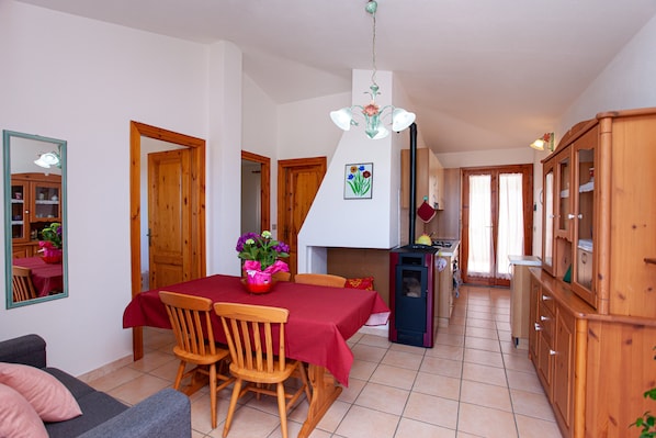 Comedor con una mesa lista para cuatro, sillas de madera, mantel rojo, candelabro y área de cocina con armarios y refrigerador.