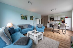 Uma sala de estar moderna com um sofá azul, uma mesa de centro branca e uma cozinha ao fundo.