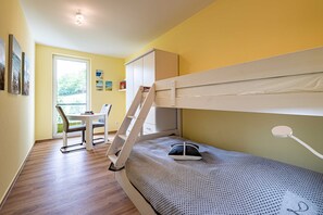 Uma cama beliche com escada, uma escrivaninha e uma cadeira em um quarto com piso de madeira e janela.