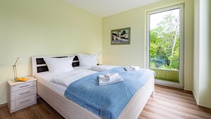 Una cama bien tendida con una manta azul, una mesita de noche con una lámpara y una ventana con vistas al verdor.