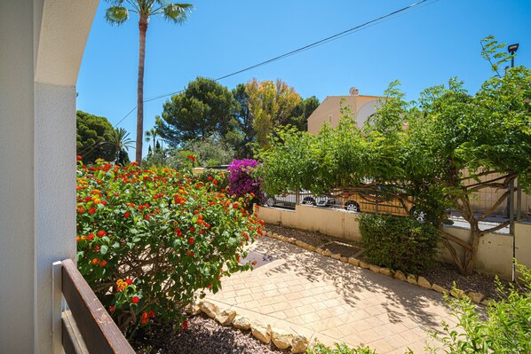 Ein Balkon mit Fliesenboden, einer Steinkante und verschiedenen Pflanzen, darunter ein Strauch mit roten Blüten und ein Baum mit grünem Laub.