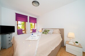 Ein Hotelzimmer mit einem Bett, einem Fernseher, einem Nachttisch mit Lampe und einem Fenster mit lila Jalousien.