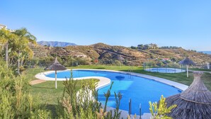 Una piscina de borde curvo, rodeada de césped y palmeras, con vistas a colinas y una estructura con techo de paja al fondo.