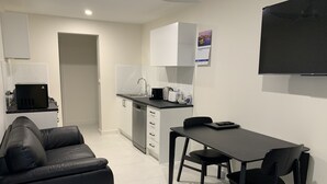 Una sala de estar moderna y minimalista con un sofá de cuero negro, una mesa de comedor con dos sillas, una cocina con armarios blancos y un televisor de pantalla plana montado en la pared.
