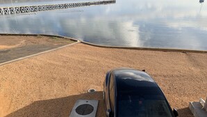 Un área de estacionamiento con un coche azul, un desagüe de hormigón y un cuerpo de agua tranquilo con un muelle al fondo.