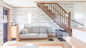 A modern living room with a wooden staircase, a grey sofa, a small round table, and a pet cage. - SEASIDE HOUSE YASUTAKETEI (Shirahama)