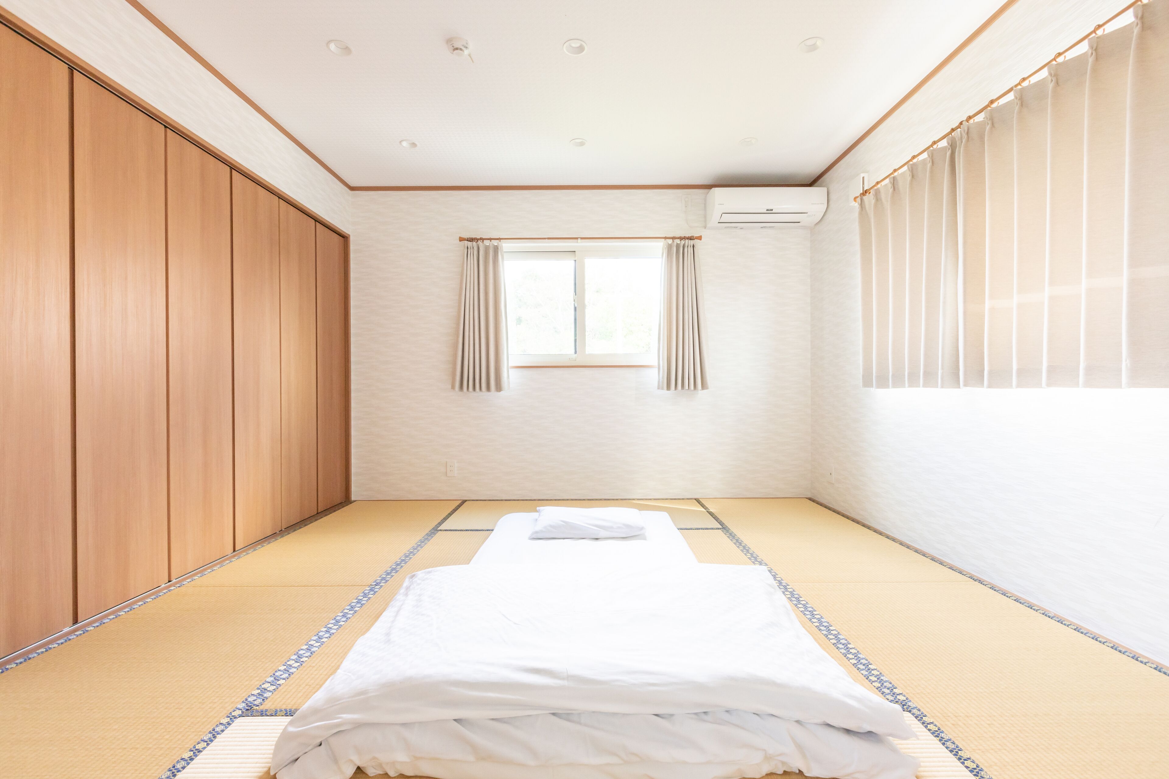 Una habitación con suelo de tatami, cama con ropa de cama blanca, puertas correderas de madera, ventana con cortinas y un equipo de aire acondicionado.