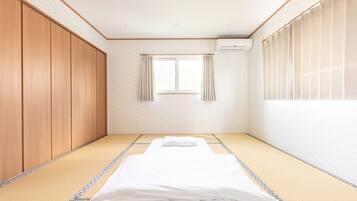 Una habitación con suelo de tatami, cama con ropa de cama blanca, puertas correderas de madera, ventana con cortinas y un equipo de aire acondicionado.