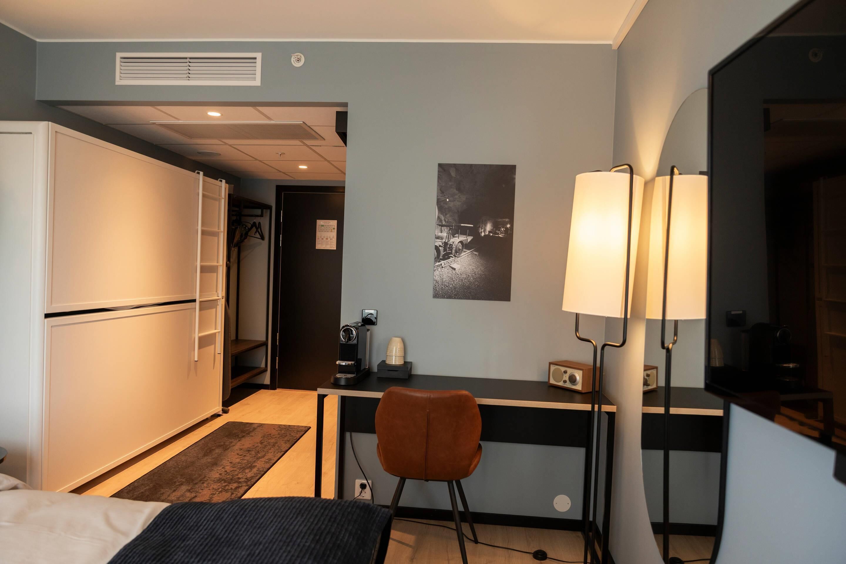 A hotel room with a bed, a desk with a chair, a lamp, a coffee machine, and a wardrobe.