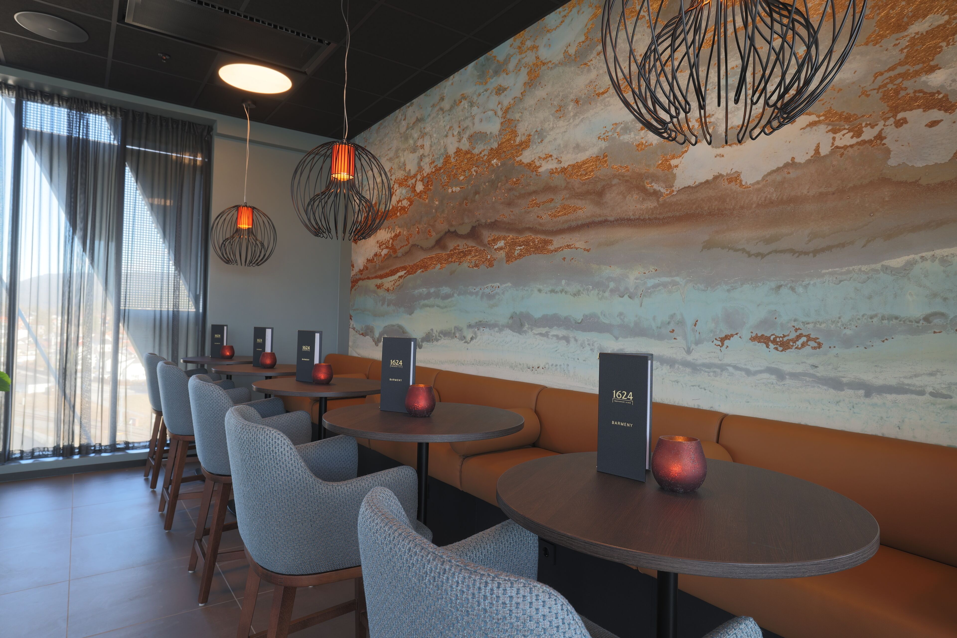 Un interior de restaurante moderno con mesas redondas, asientos acolchados, iluminación colgante y un mural texturizado en la pared.