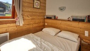 A wooden cabin room with a bed, a window with curtains, a wall-mounted shelf with books, and a framed picture. - Mountain apartment, sleeps 6, south balcony (Morillon 1100)