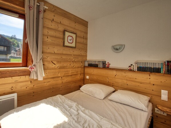 Habitació de cabana de fusta amb un llit, una finestra amb cortines, una estanteta penjada amb llibres i un quadre amb marc.