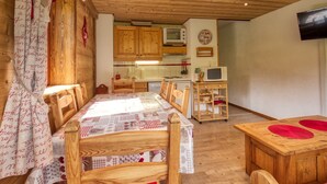 A cozy room with wooden furniture, a dining table, a kitchen area with wooden cabinets, and a television mounted on the wall. - Mountain apartment, sleeps 6, south balcony (Morillon 1100)