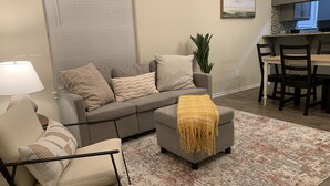 Ein Wohnzimmer mit einer Couch, einem Sessel, einem kleinen Hocker, einer Lampe, einer Pflanze und einem Essbereich mit Tisch und Stühlen.