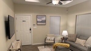 Ein Wohnzimmer mit grauem Sofa, einem kleinen Sessel, einem Couchtisch, einem an der Wand montierten Fernseher, einem Deckenventilator und einem Fenster mit Jalousien.