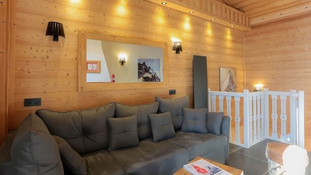 A cozy wooden cabin interior with a sectional sofa, a wooden ceiling with recessed lighting, a white railing, and a framed picture on the wall.