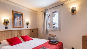 A bedroom with a wooden headboard, a large bed with red and white bedding, two wall-mounted lamps, a framed picture, and a window with a view of a building.