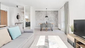 Ein modernes Wohnzimmer mit einem grauen Sofa, einem Esstisch mit Stühlen, einem Fernseher und einem großen Fenster mit Vorhängen.