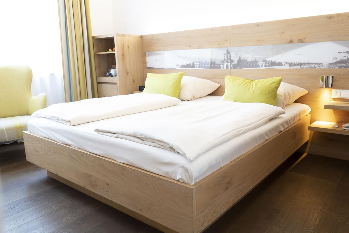 A double bed with a wooden frame, two pillows, a bedside table with a lamp, and a wall-mounted shelf.
