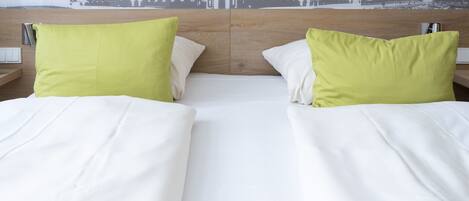 Una cama doble con ropa blanca de cama y dos almohadas, una verde y otra blanca, en una habitación con cabecera de madera y un mural con vista a la ciudad.