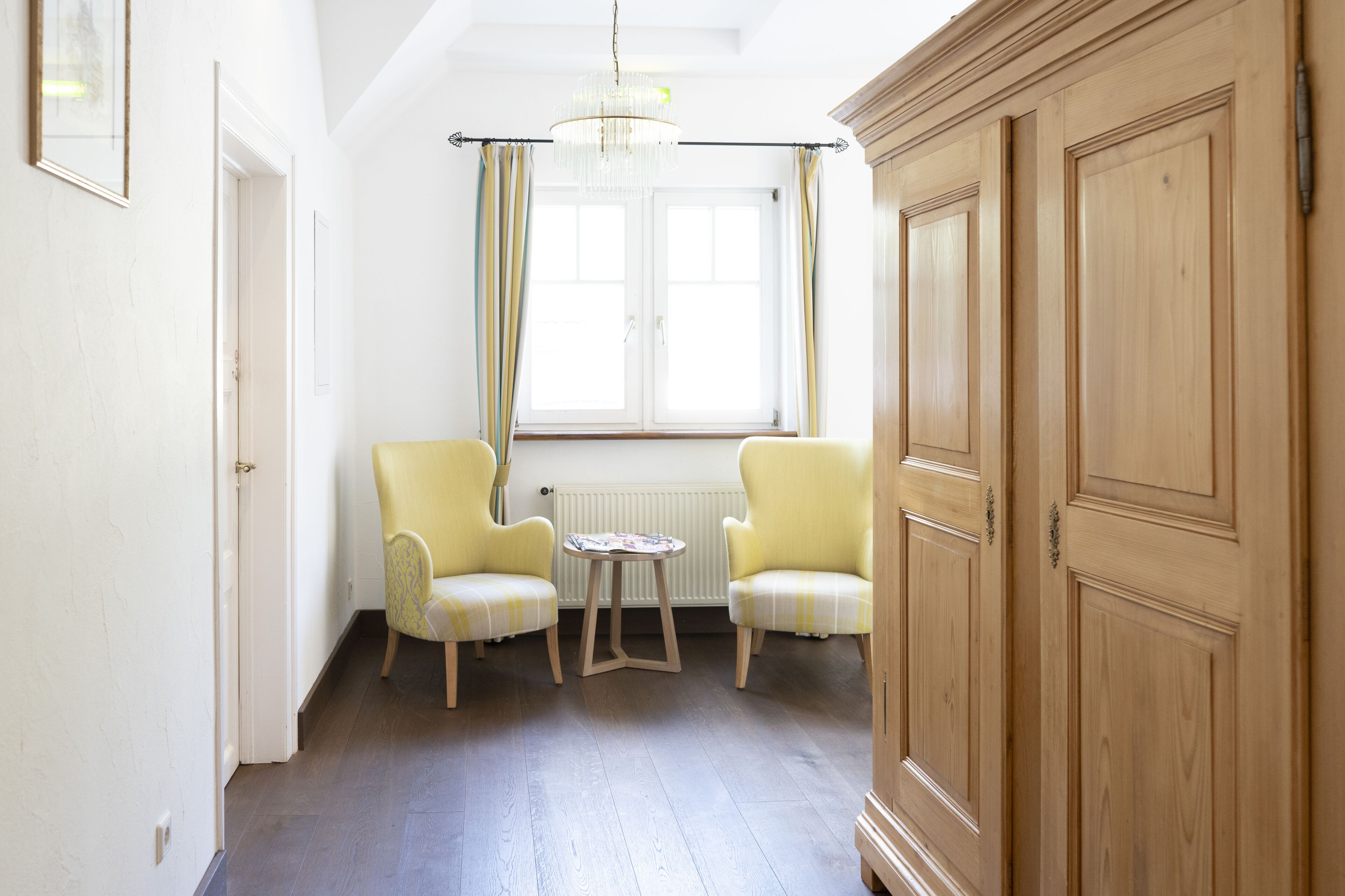 Una habitación con dos sillones amarillos, una mesa redonda pequeña, un armario de madera y una ventana con cortinas.