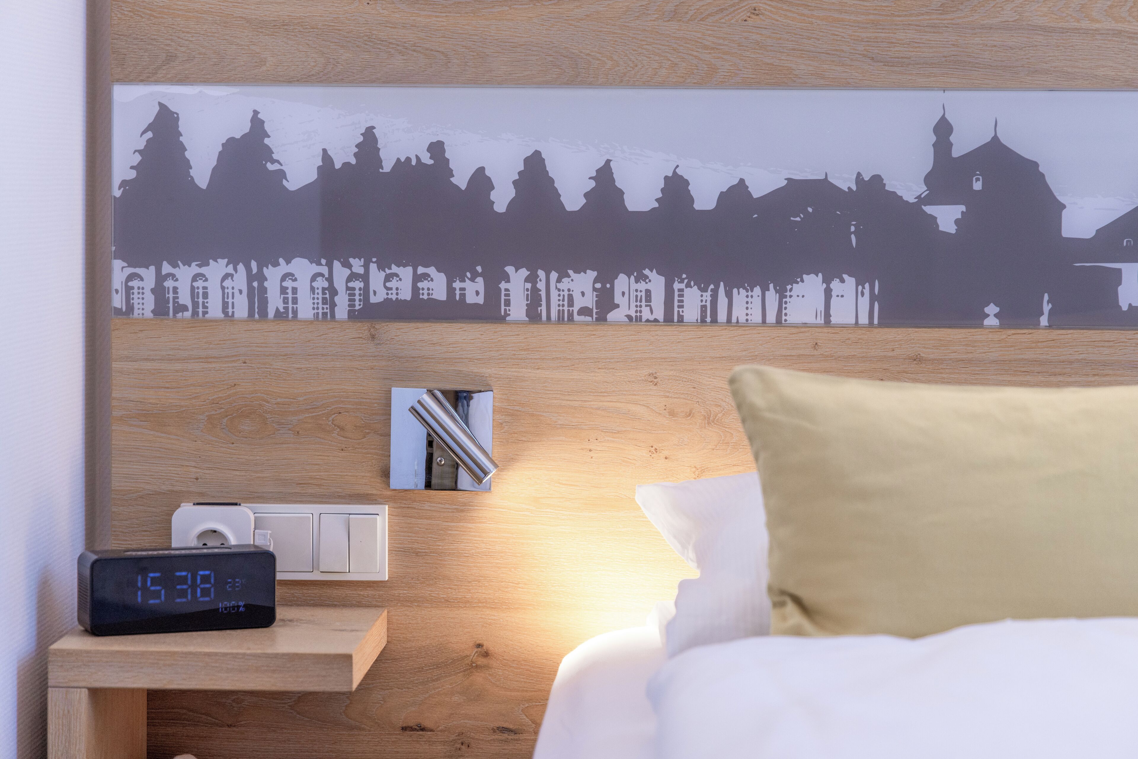 A hotel room with a bed, a bedside table with a digital clock, a wall-mounted air conditioner, and a framed picture of a cityscape.