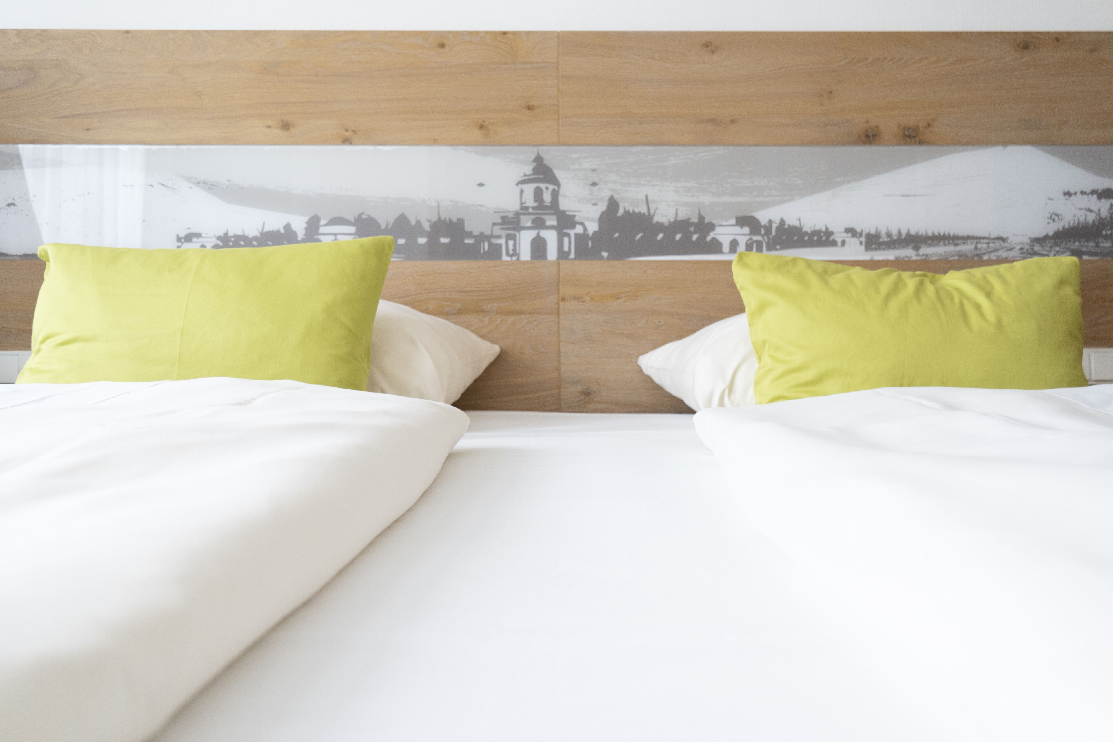 Ein Bett mit weißer Bettwäsche und zwei leuchtend grünen Kissen, vor einem Holzkopfteil mit Stadtansicht-Wandgemälde.