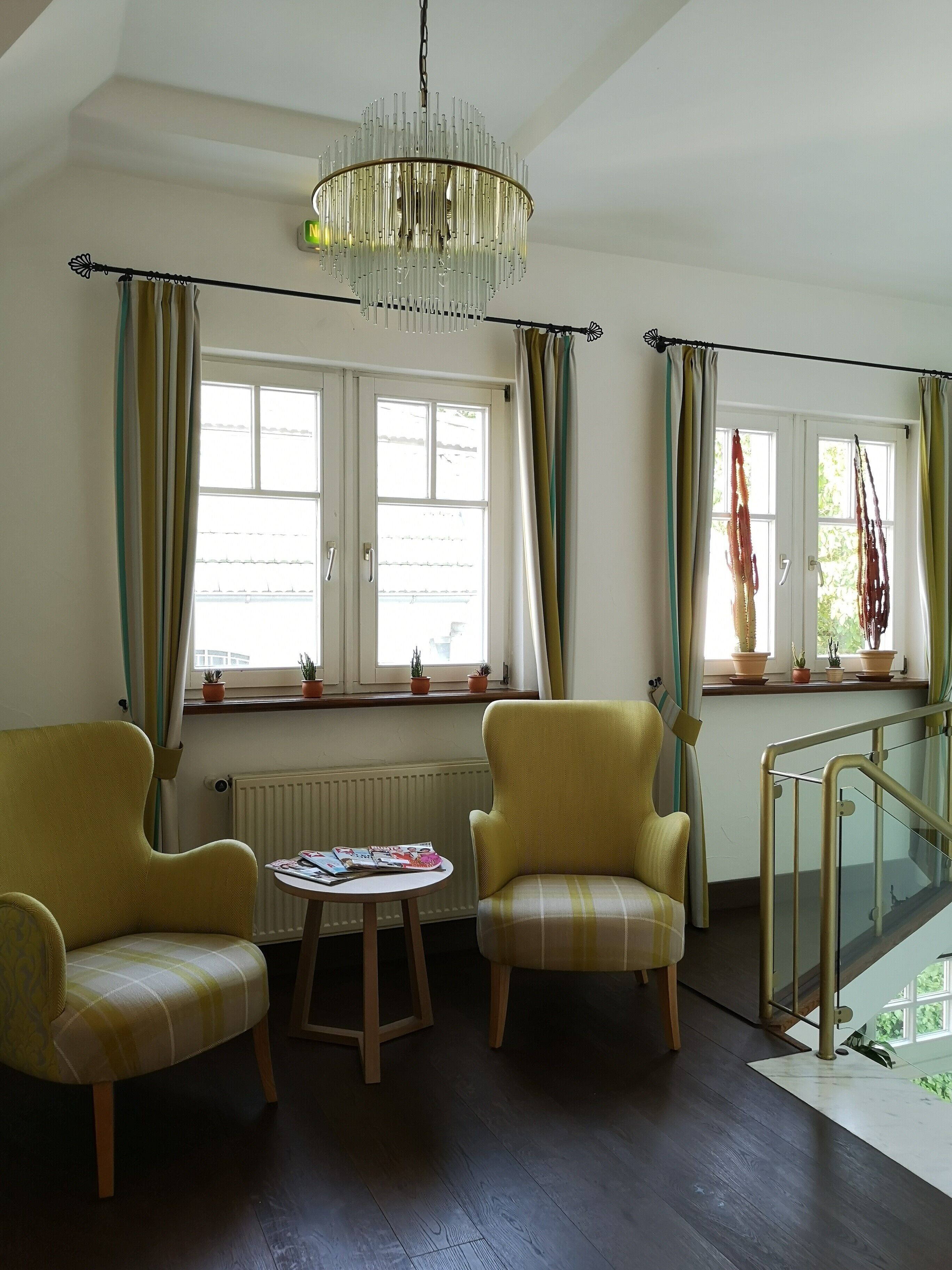 A room with two yellow armchairs, a small round table with magazines, a chandelier, and large windows with curtains.