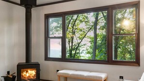 A room with a wood-burning stove, a bench, and a large window with a view of trees.