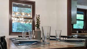 A dining table set with clear glasses, plates, and cutlery, with a vase of flowers and a string of lights in the background.
