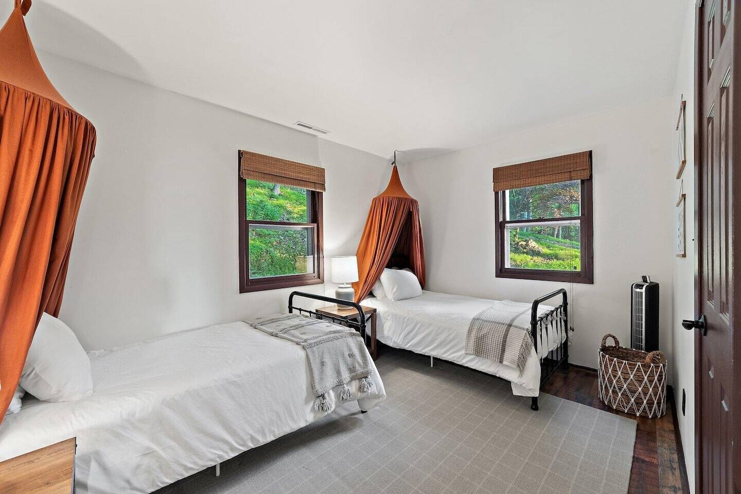 Quarto com duas camas de solteiro, mesa de cabeceira com luminária, janela com vista para área verde e porta com um cesto no chão.