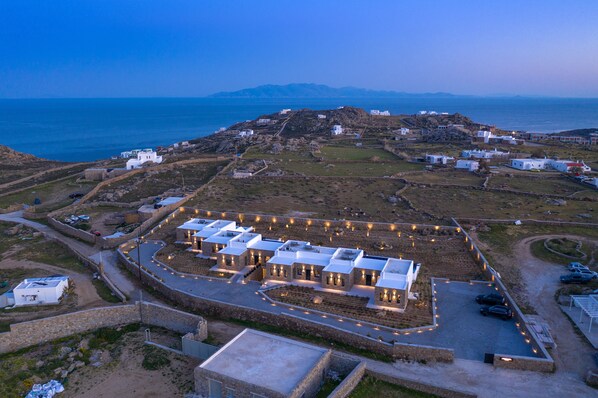 An aerial view of a coastal area with a series of white buildings, a parking lot, and a stone wall surrounding the property. - Nimbus Eden (Mykonos)
