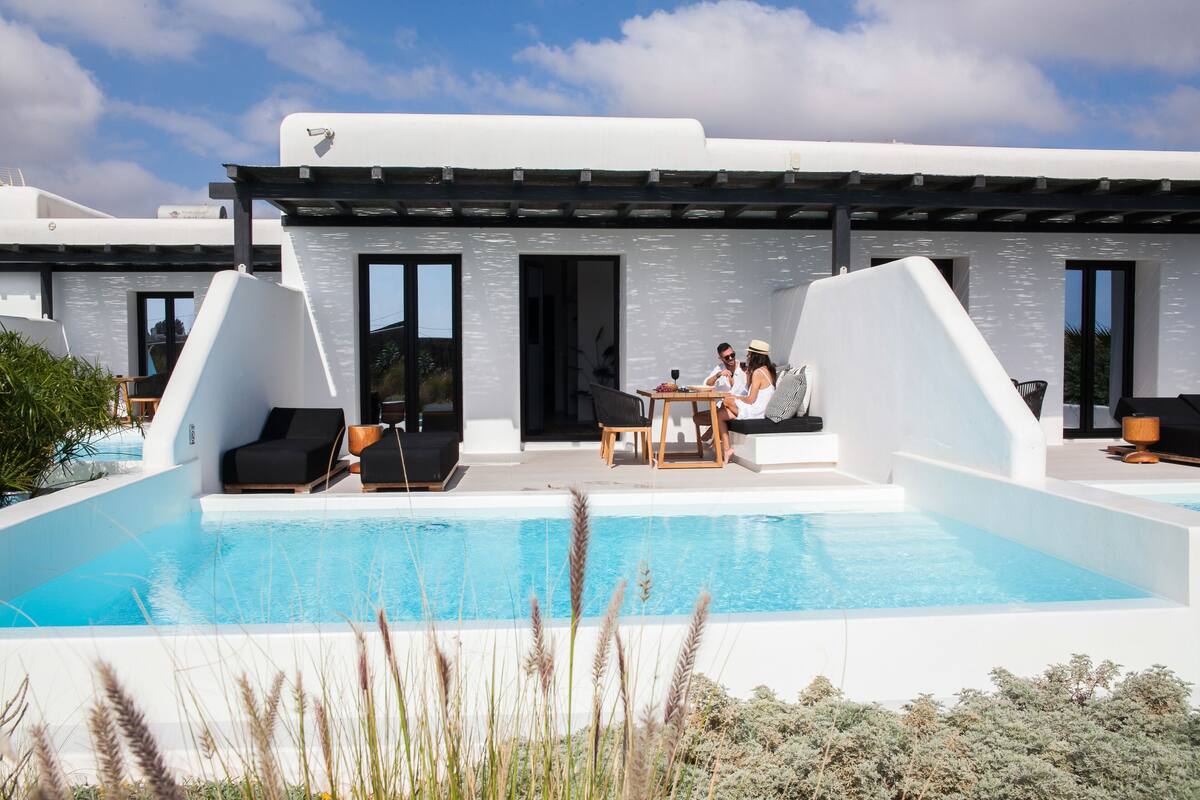 Un área de piscina exterior moderna con un edificio blanco, mobiliario negro y un cielo azul despejado.