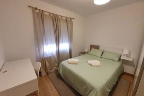 Una cama individual con una colcha verde, una mesita de noche con una lámpara, una silla y un escritorio con un hueco en el centro.