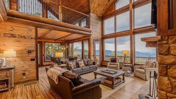 A spacious living room with a large fireplace, wooden flooring, and a balcony with a view of the mountains. - Experience breathtaking mountain views at Sequoyah Sunset (Bryson City)