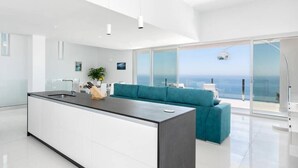Une cuisine moderne avec un îlot central, un canapé de couleur turquoise et une grande porte-fenêtre coulissante donnant sur un balcon avec vue sur l’océan.