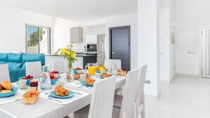 Une pièce moderne et bien éclairée, avec une table à manger dressée pour le petit-déjeuner, un canapé bleu et un coin cuisine à l’arrière-plan.