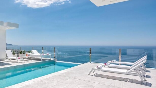 Une piscine à débordement moderne, avec une eau d’un bleu cristallin, des chaises longues blanches et une vue sur l’océan.