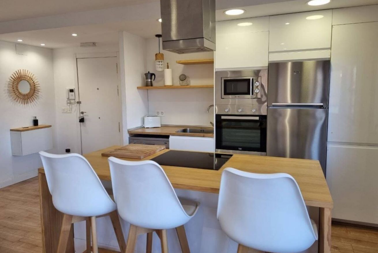 Een moderne keuken met een centraal eiland, witte stoelen, roestvrijstalen apparatuur en een houten eettafel.