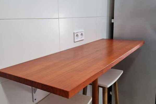 A wooden table with a white base, positioned against a white tiled wall with a power outlet.