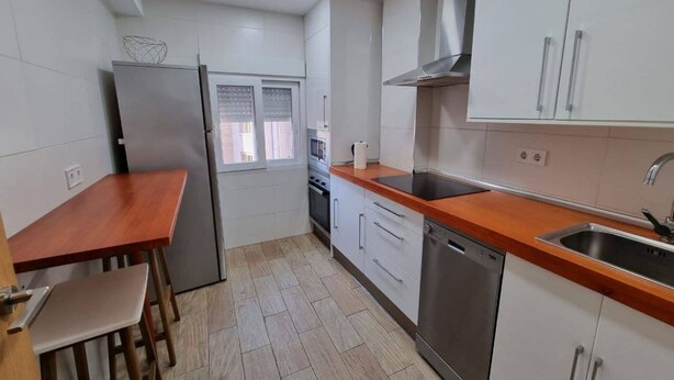A compact kitchen with white cabinets, a stainless steel refrigerator, a built-in oven, a dishwasher, and a window with blinds. - 103A By R2R 2-Bedroom Apartment (Gijón)