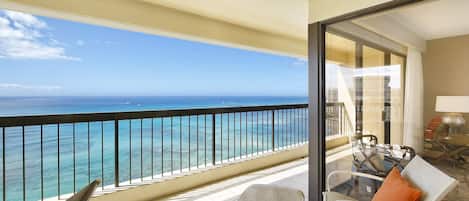 A balcony with a view of the ocean, a table with chairs, and a glass of drink.