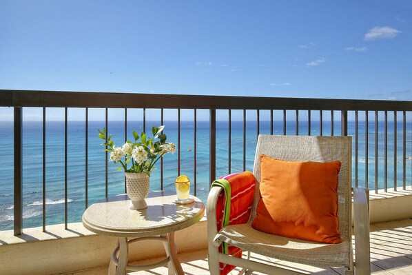 Ein Balkon mit Blick auf den Ozean, ein Stuhl mit Kissen, ein kleiner Tisch mit Blumenvase und ein Glas mit Getränk.