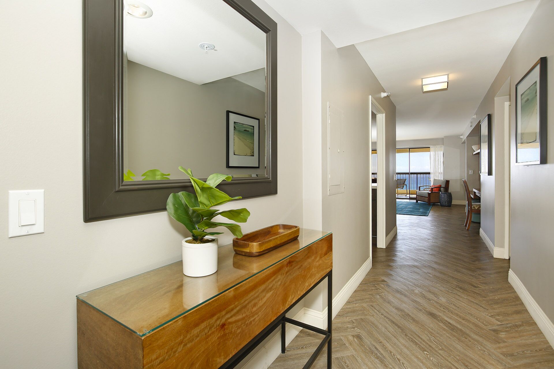 A hallway with a wooden floor, a glass and wood vanity with a plant, a large mirror, and a framed picture on the wall.