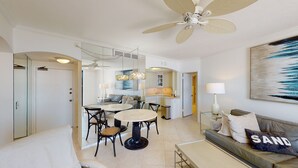 Ein modernes Wohnzimmer mit einem runden Esstisch, schwarzen Stühlen, einer Couch mit einem 'SAND'-Kissen, einem Deckenventilator und einer Küche im Hintergrund.