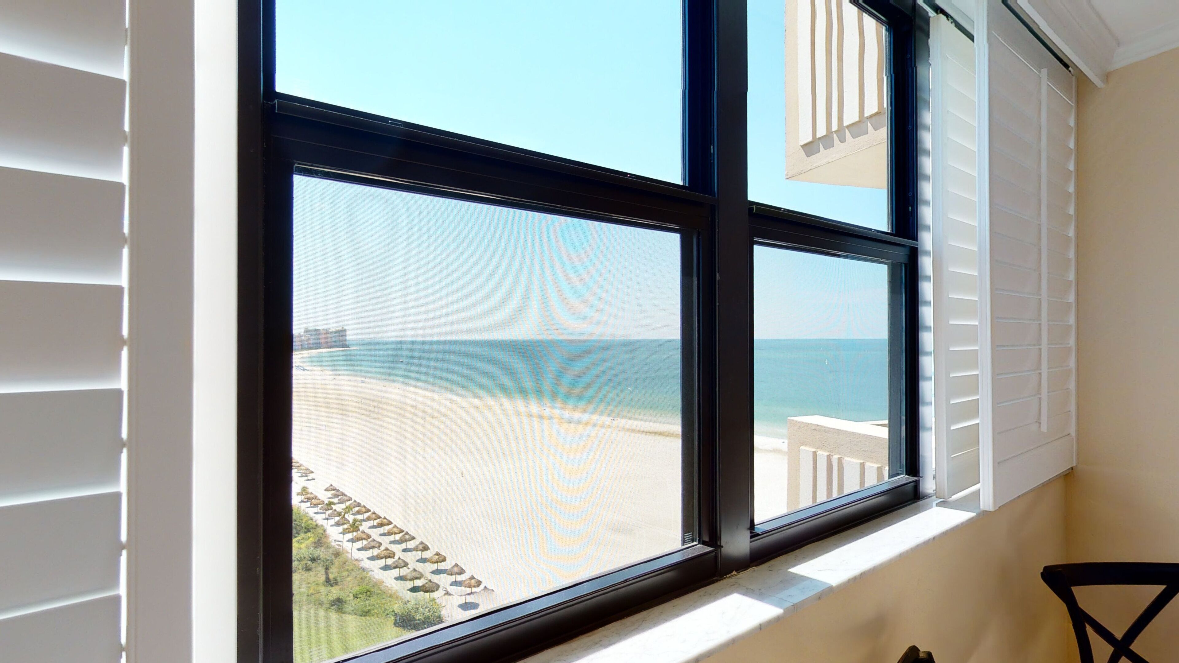 A window with a view of a beach, a building with white shutters, and a chair.