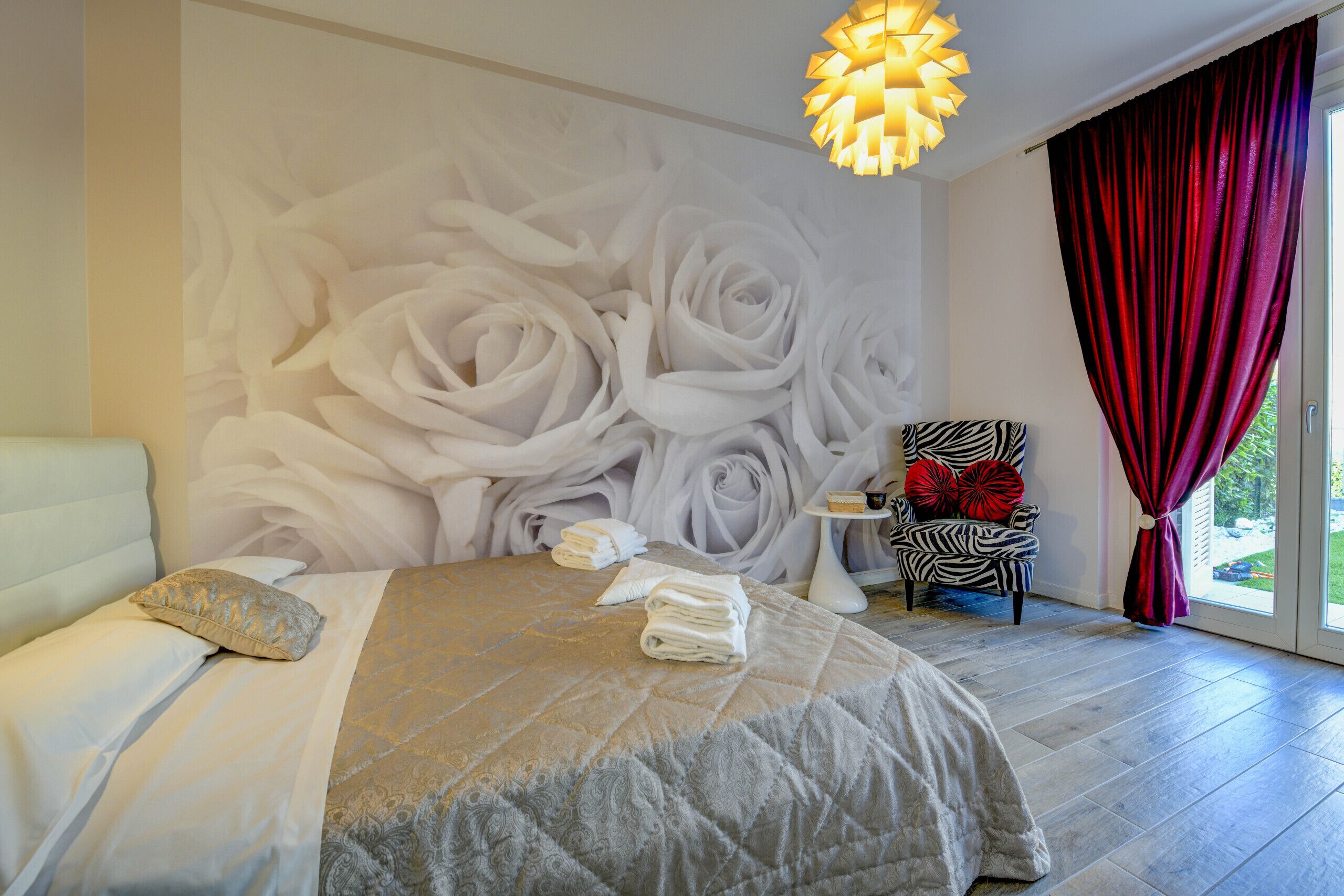 A bedroom with a large wall mural of white roses, a bed with a grey and white duvet, a zebra-patterned chair, and a red curtain.